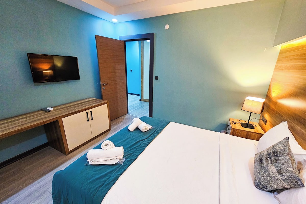 A hotel room with a bed, a TV mounted on the wall, a wooden wardrobe, a nightstand with a lamp, and a door leading to another room.