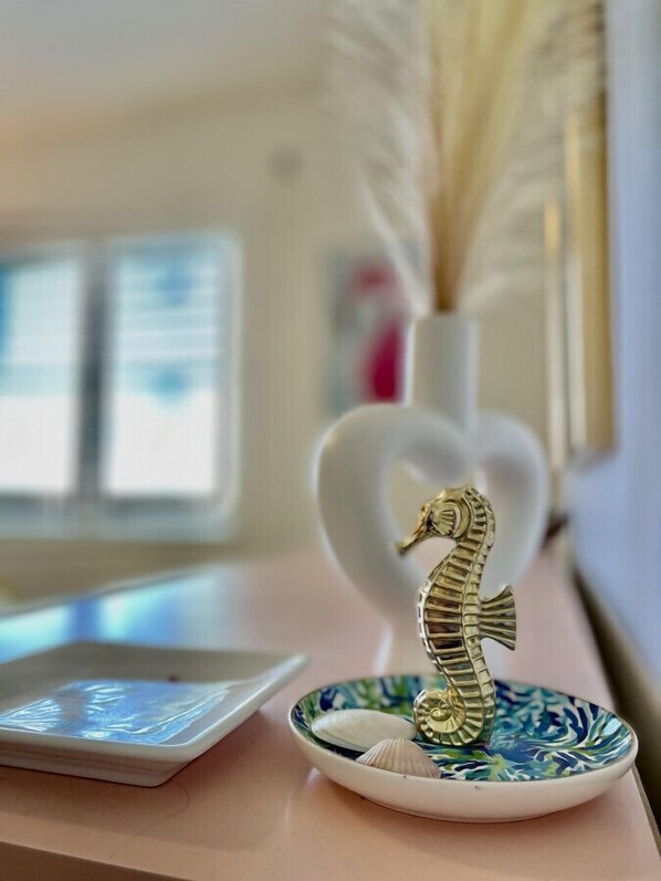 A seahorse figurine on a decorative plate with shells, a white vase with feathers, and a window in the background.