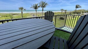 A deck with a round table and two Adirondack chairs, overlooking a beach with palm trees and the ocean. - Sea Oats 3BR-Direct Beach Access with Pool (Cocoa Beach)