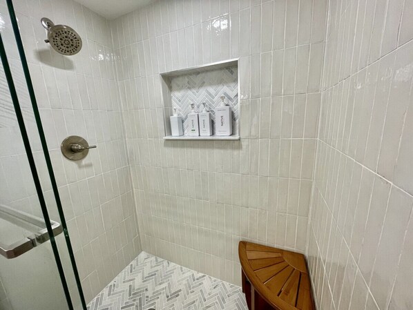 A shower with a glass door, a wooden bench, and a shelf with toiletries. - Sea Oats 3BR-Direct Beach Access with Pool (Cocoa Beach)