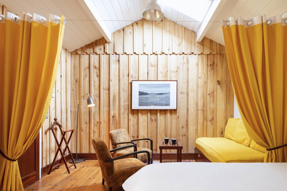 Une pièce avec un mur lambrissé en bois, un tableau encadré, un canapé jaune, un fauteuil marron et un lit.