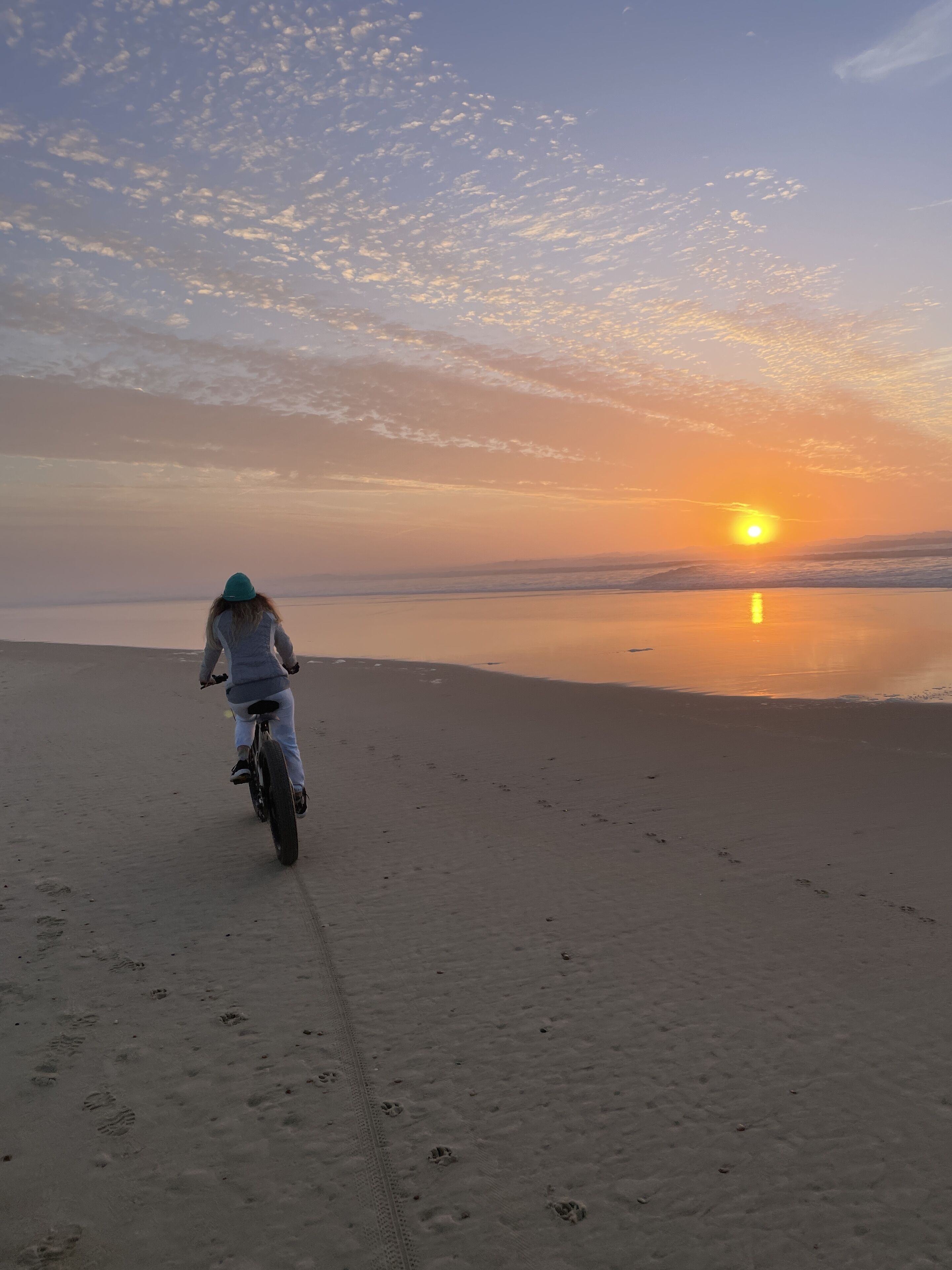 Eine Person fährt mit einem Fahrrad über einen sandigen Strand, im Hintergrund ein Sonnenuntergang.