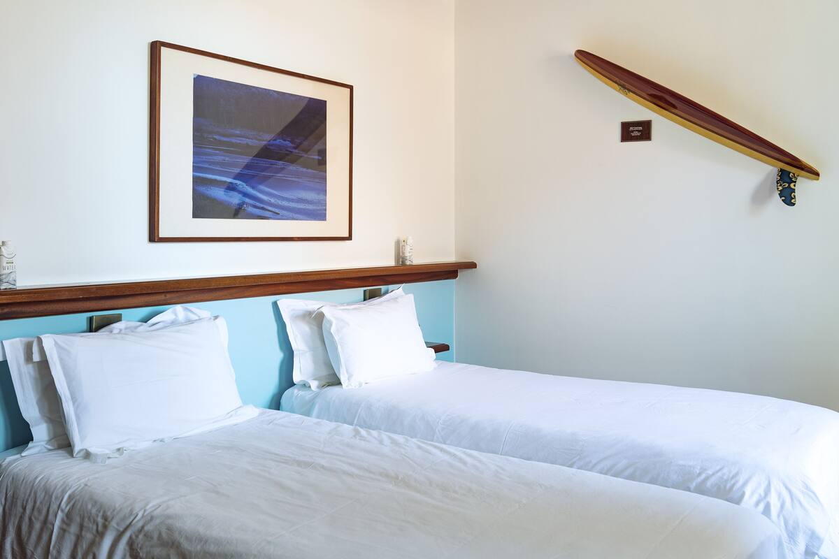 Une chambre d’hôtel avec deux lits, un tableau encadré au mur et une planche de surf accrochée au mur.