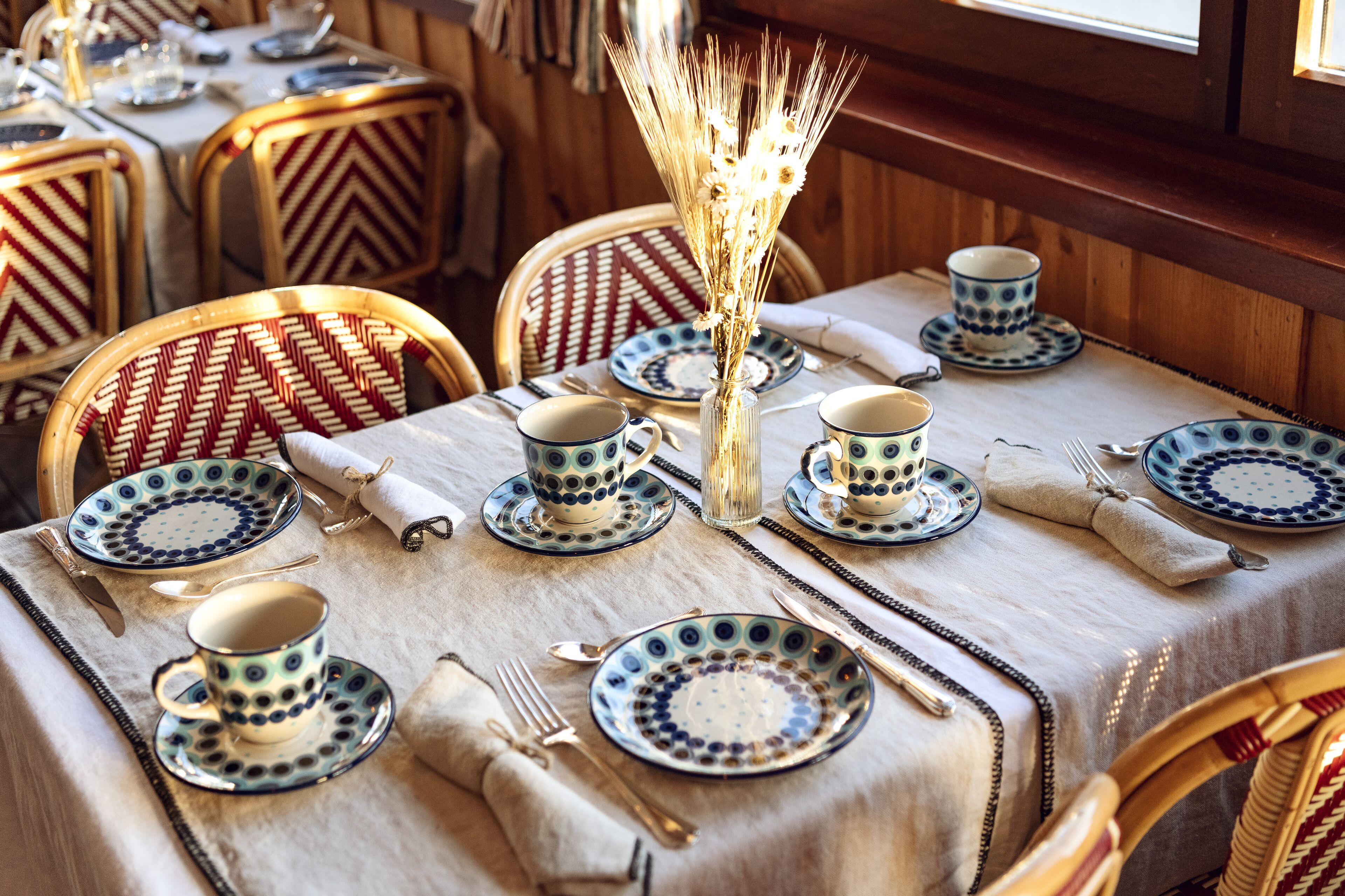 A dining table set with blue and white patterned plates, cups, and cutlery, with a vase of dried flowers in the center.