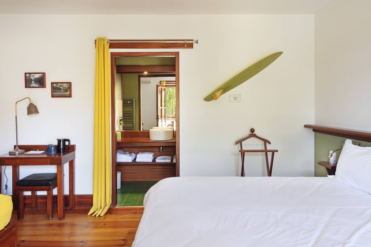 Une chambre d’hôtel avec un lit, un bureau doté d’une lampe, une chaise, une planche de surf accrochée au mur et une salle de bain visible à travers une porte ouverte.