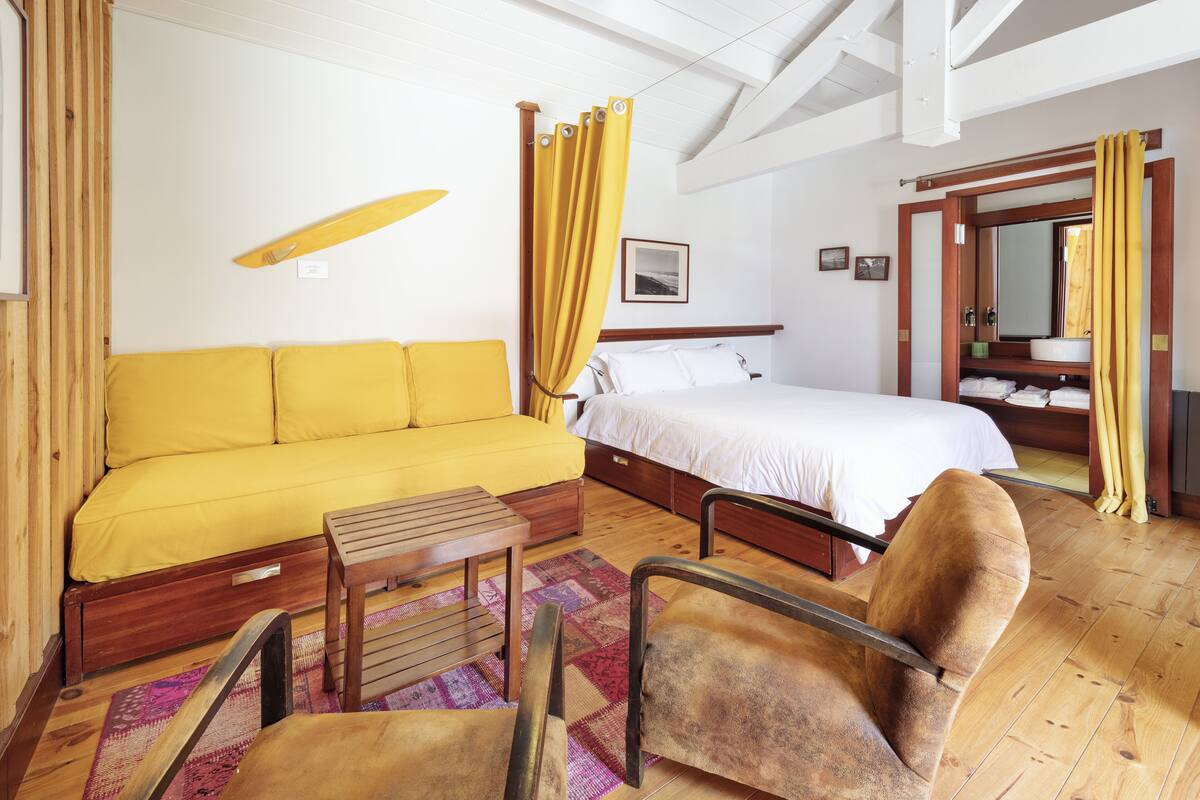 Une pièce avec un canapé jaune, un lit, une chaise en bois et une petite table.