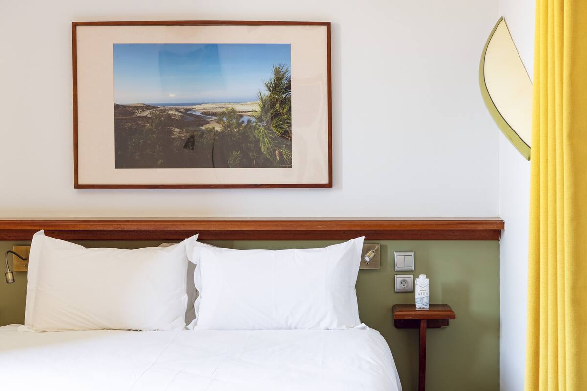 Un lit bien fait, avec du linge de lit blanc, un tableau représentant un paysage accroché au mur et une table de chevet sur laquelle se trouve une bouteille d’eau.