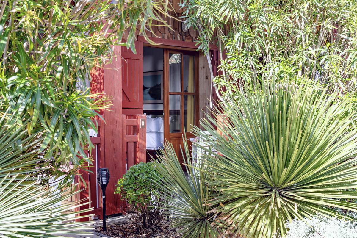 Un bâtiment en bois peint en rouge, doté d’une porte et de fenêtres, et entouré de différentes plantes.