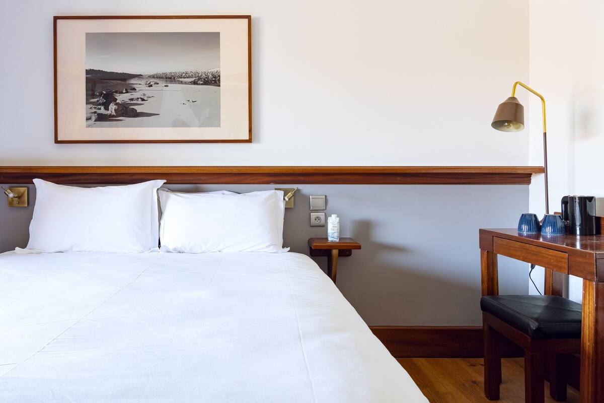 Une chambre d’hôtel avec un lit bien fait, une photographie encadrée accrochée au mur, une table de chevet avec une lampe et un bureau en bois avec une chaise.