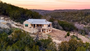 Una casa moderna de dos pisos con un balcón grande, rodeada de un bosque denso y colinas.