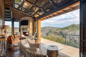 A spacious balcony with wicker furniture, a stone fireplace, and a view of the surrounding landscape. - Direct Access to River & Scenic Views | Sotol by AvantStay (Dripping Springs)