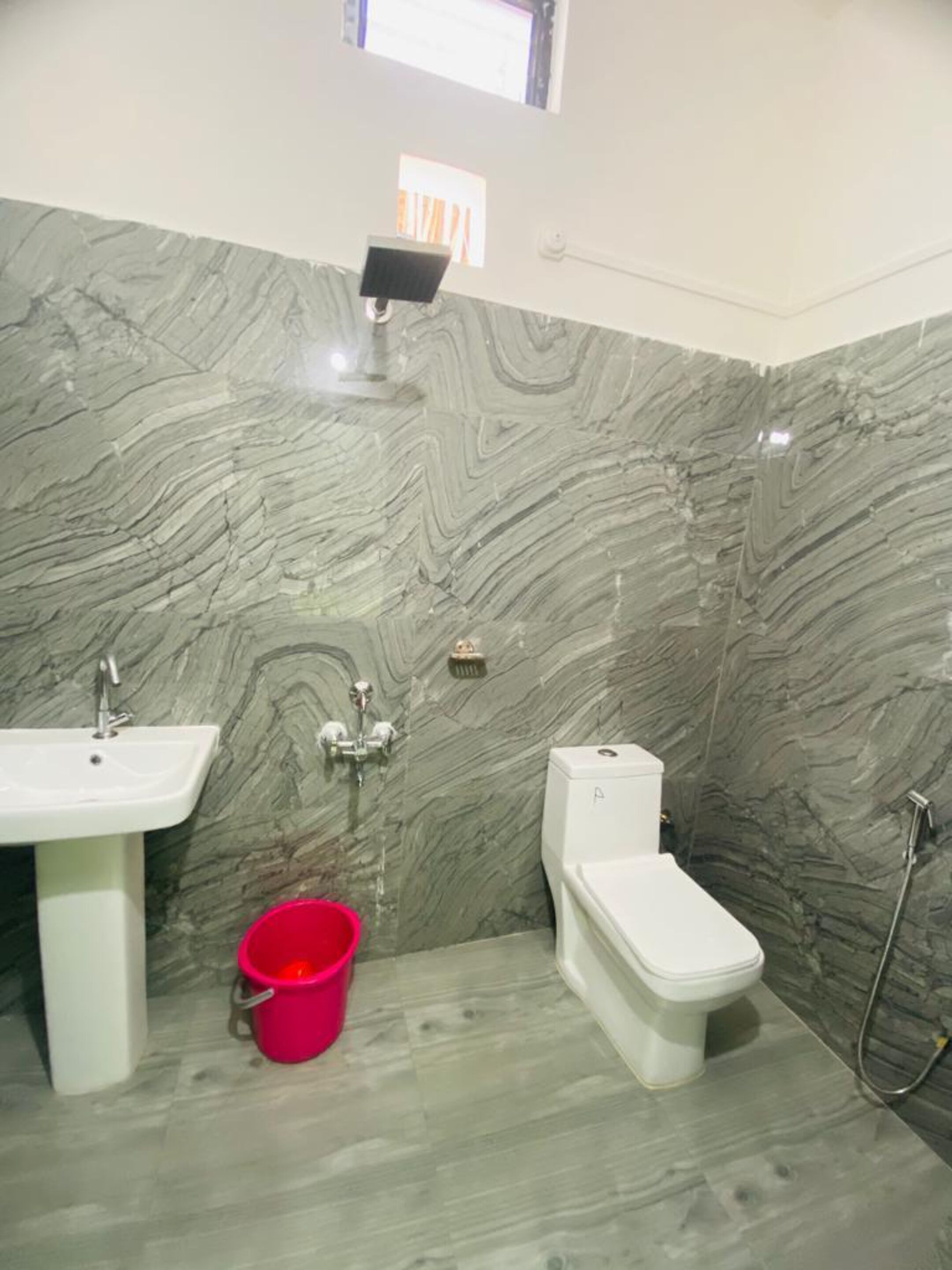 Een badkamer met een marmeren wand, een wastafel, een toilet en een rode emmer op de vloer.