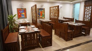 A restaurant with wooden furniture, tables set with plates and cutlery, and decorative wall art. - Shri Nilayam Kutir (Varanasi)