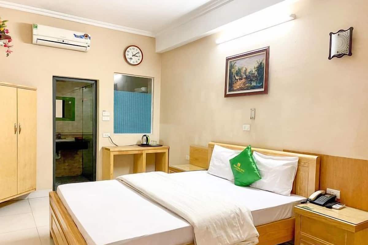 A hotel room with a bed, bedside table, and a desk with a chair. There is a clock on the wall and an air conditioning unit above the desk.