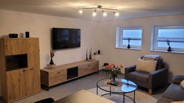 A modern living room with a wooden cabinet, a flat-screen TV, a sofa, a coffee table, and a window with a view. - Ferienwohnung Hereinspaziert (Wilhelmshaven)