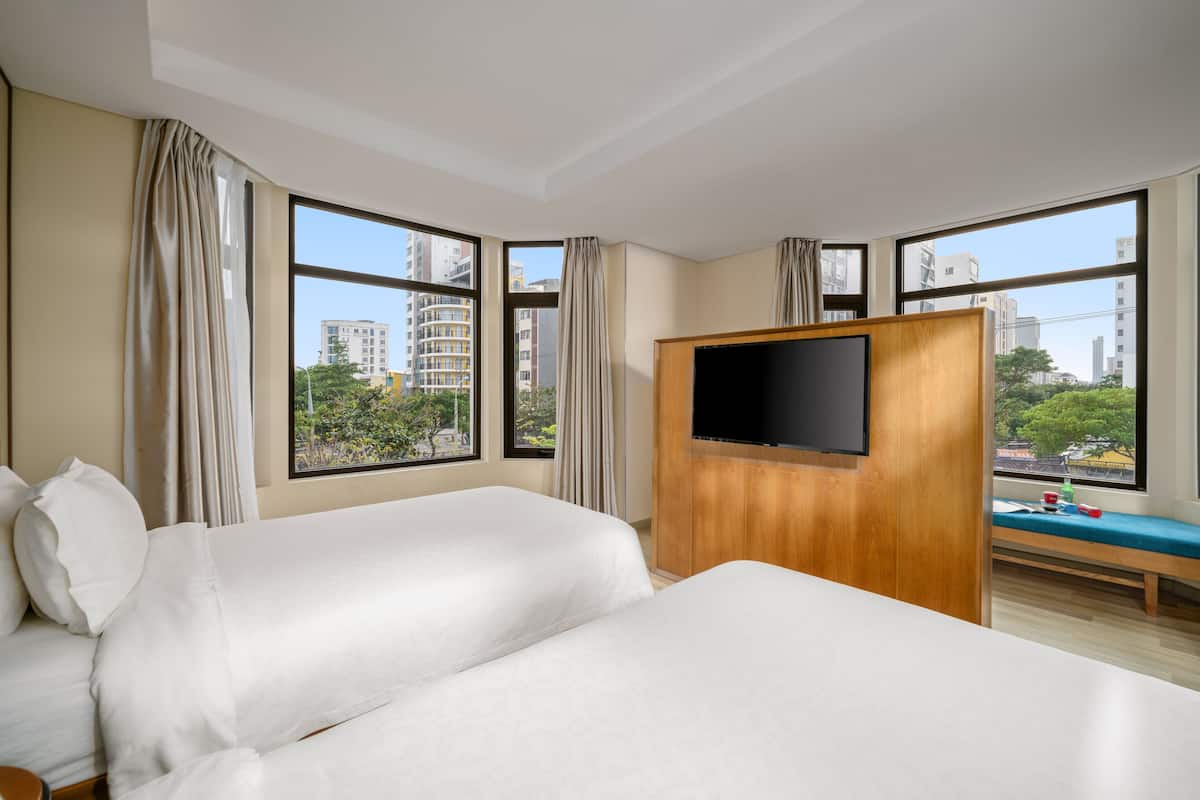 Habitación de hotel con dos camas, televisor, un banco con cojín y ventanas amplias con vista a la ciudad.