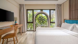 Habitación de hotel con una cama grande, un escritorio con una computadora portátil, un televisor y vistas a zonas verdes a través de una ventana.