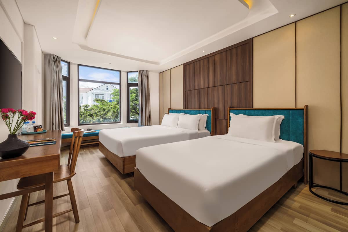 Habitación de hotel con dos camas, un escritorio de madera, una silla y un amplio ventanal con vistas a zonas verdes.