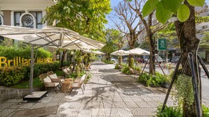 Un área de café al aire libre con mesas y sillas bajo sombrillas, rodeada de follaje y árboles.