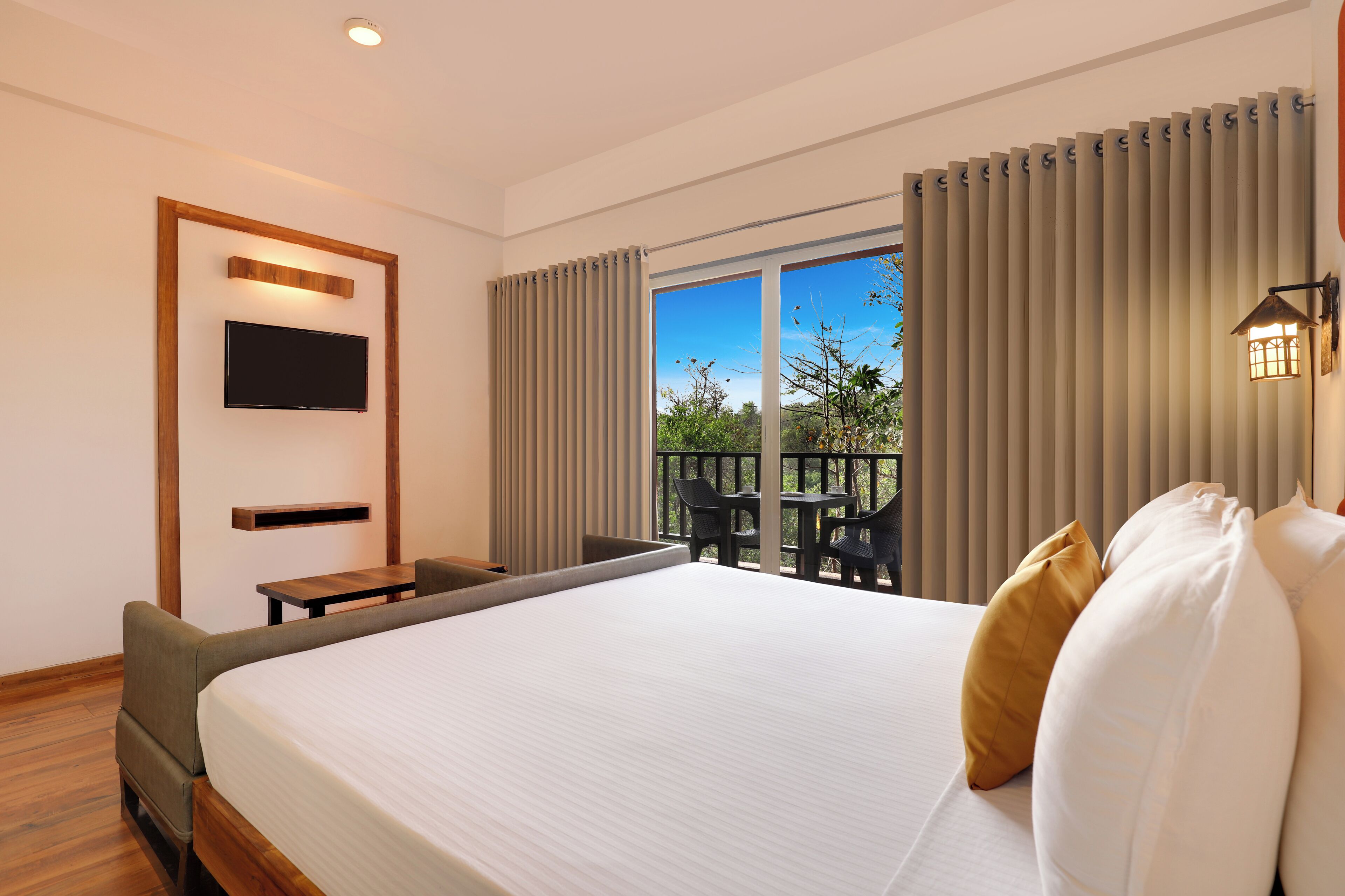 A hotel room with a large bed, a TV mounted on the wall, a balcony with chairs, and a view of the outdoors.