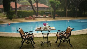 A poolside area with a table set for two, surrounded by ornate benches, and a view of a residential area in the background. - Sariska Tiger Heaven Resort (Thanagazi)