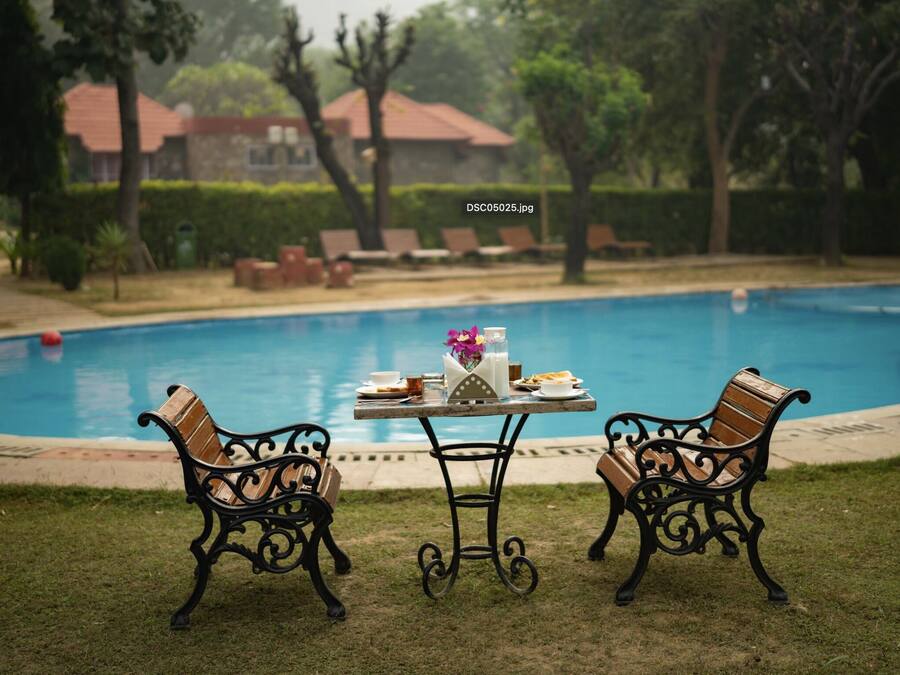 Un área junto a la piscina con una mesa dispuesta para dos personas, rodeada de bancos ornamentados, y vista a una zona residencial al fondo.