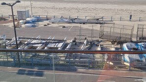 A beach view from a balcony with a chair and a table, overlooking a parking area with boats and wind turbines.