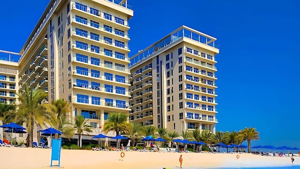 A beachfront hotel with multiple floors, clear blue water, and palm trees. - UAEVACAY (Ras Al Khaimah)