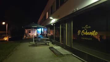 A nighttime view of a building with a sign reading "El Cubanito" and a website address. There are outdoor seating areas with tables and benches, and a covered area with a canopy.