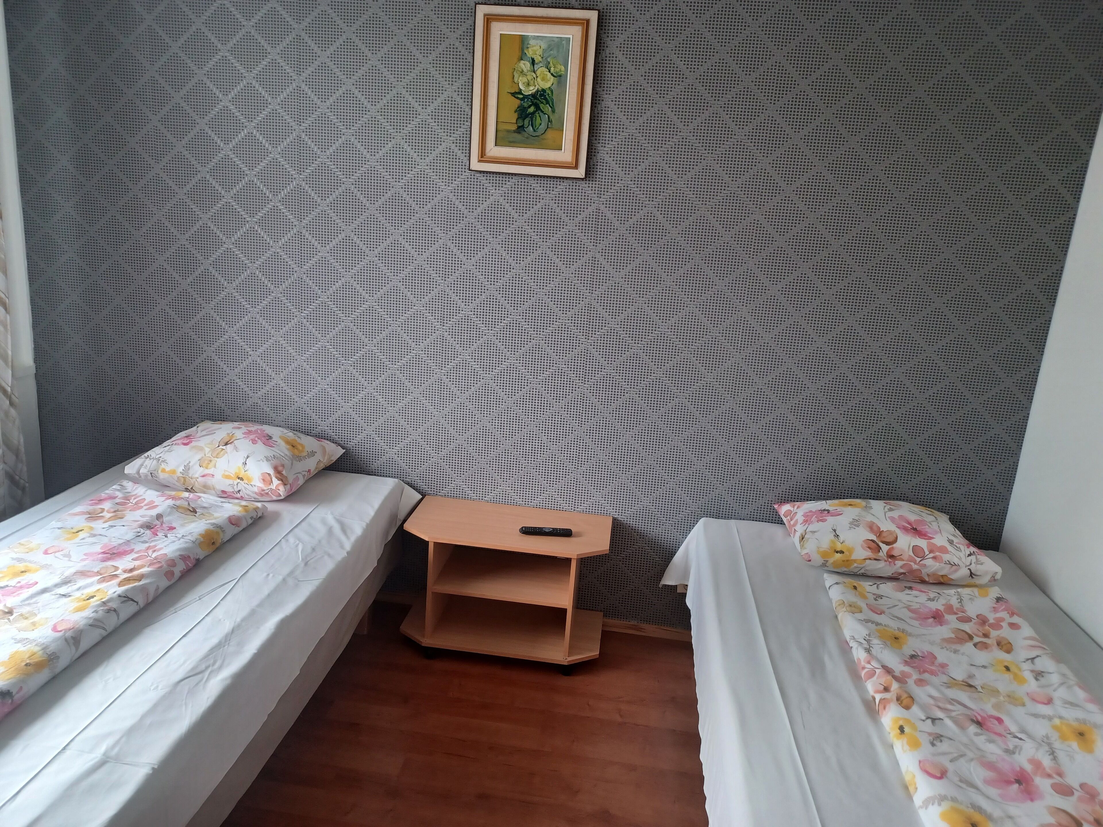 A room with two single beds, each with floral-patterned bedding. There is a small wooden nightstand between the beds, and a framed picture hangs on the wall above.