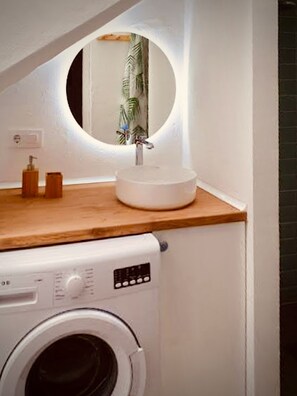 Un espace buanderie compact comprenant un lave-linge, un évier avec un miroir et une petite étagère pour les articles de toilette.