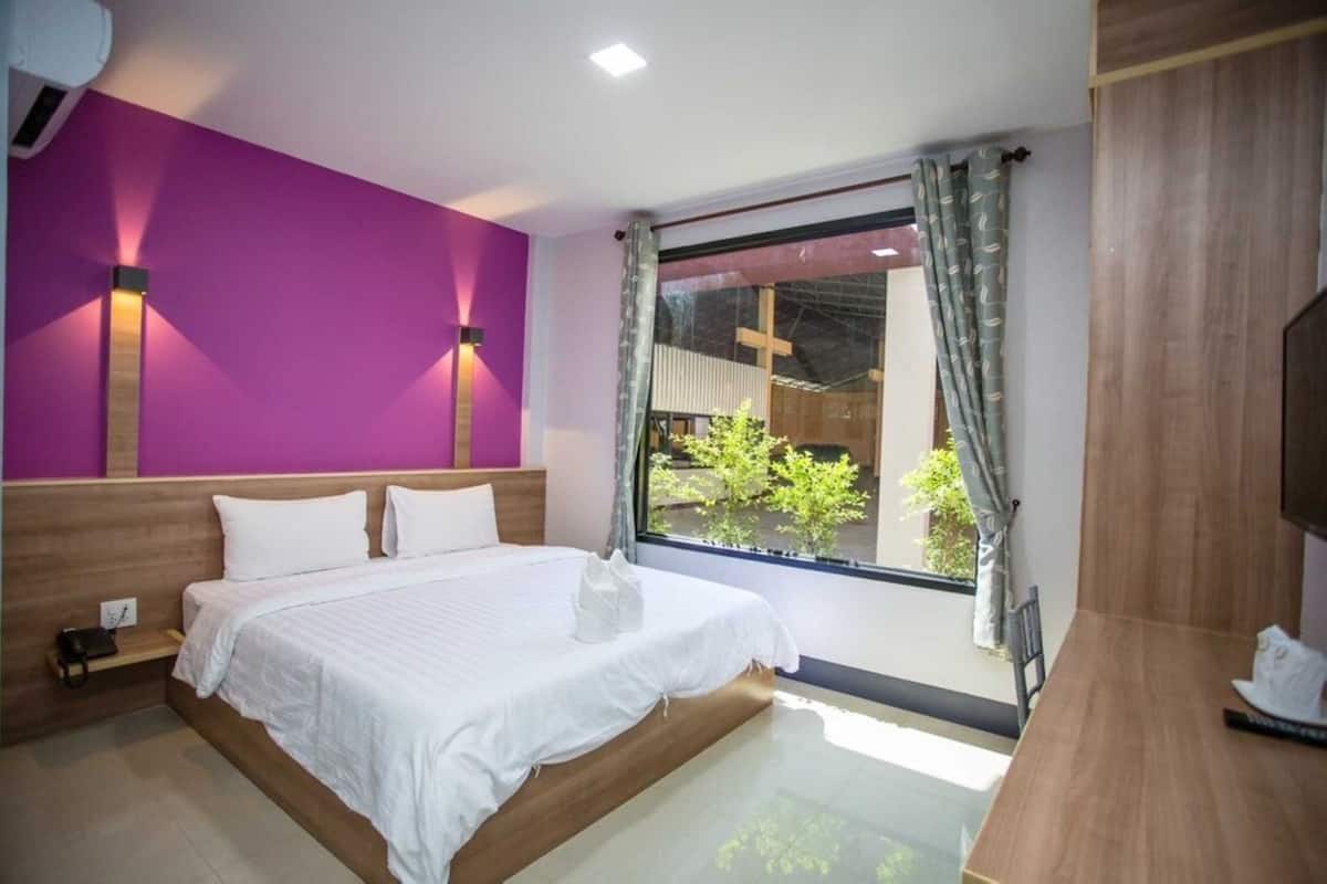 Une chambre d’hôtel moderne avec un grand lit, une tête de lit en bois, une fenêtre donnant sur un espace verdoyant et une petite table avec une chaise.