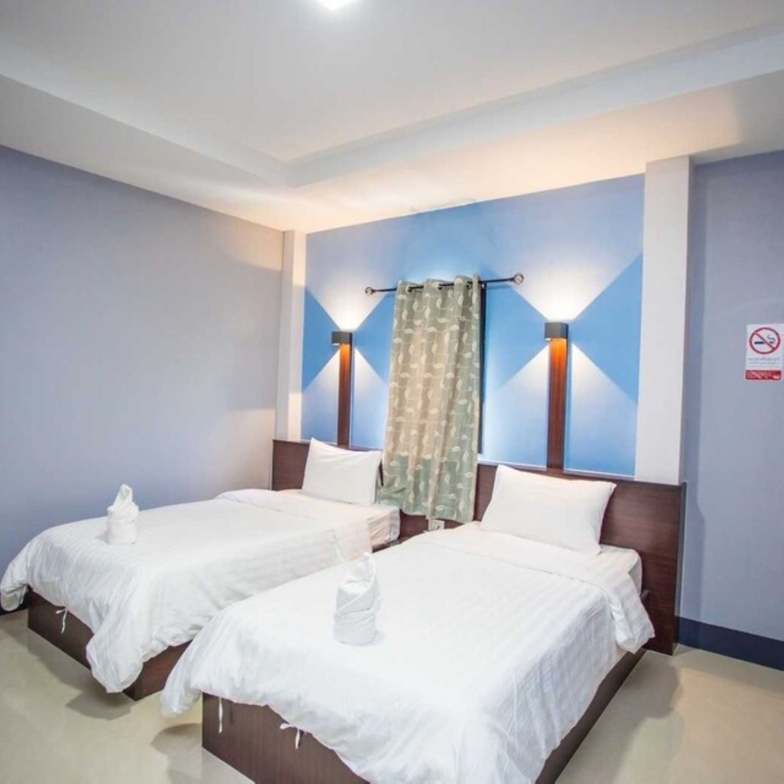 A hotel room with two single beds, a nightstand, a wall-mounted lamp, and a curtain.