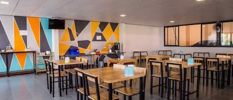 Un área de comedor moderna con varias mesas y sillas de madera, una barra con dispensador de bebidas y una pared con estampado geométrico.
