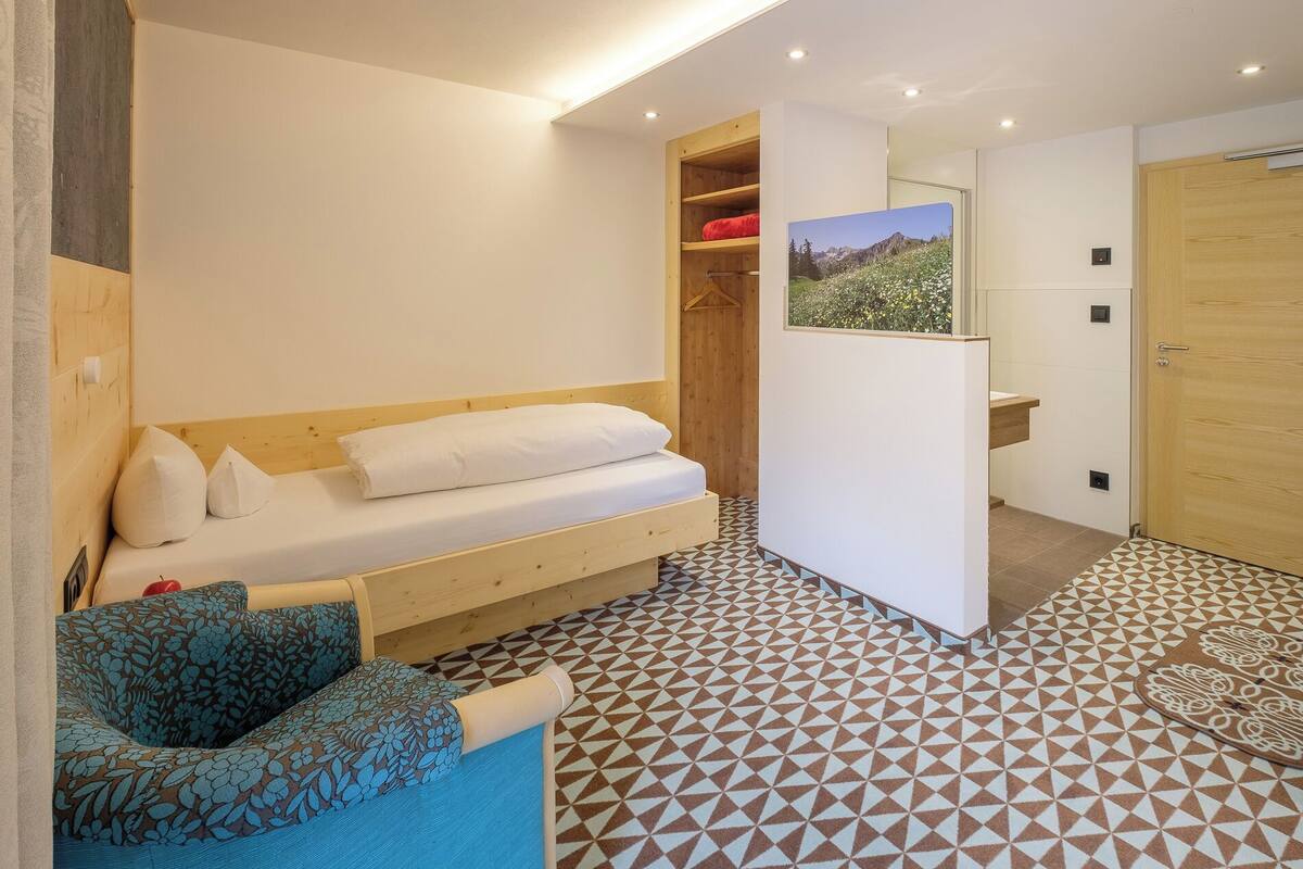 A bedroom with a wooden bed, a blue patterned sofa, a geometric patterned floor, a wooden wardrobe, and a framed picture of a mountain landscape.