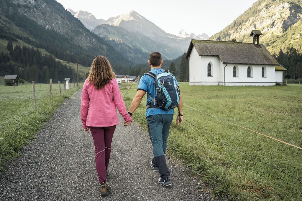 A couple walking on a gravel path towards a white building with a bell tower, surrounded by mountains.