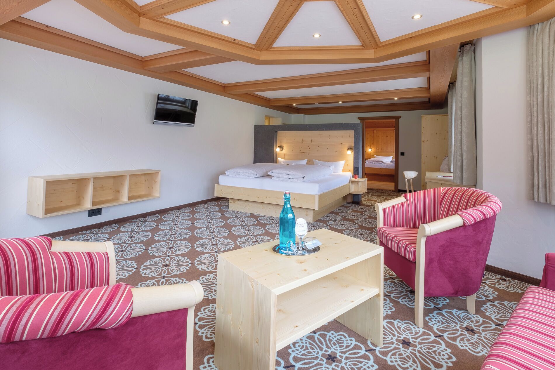 A hotel room with a wooden ceiling, a bed with white linens, a wooden nightstand, a chair, and a small table with a bottle and glasses.