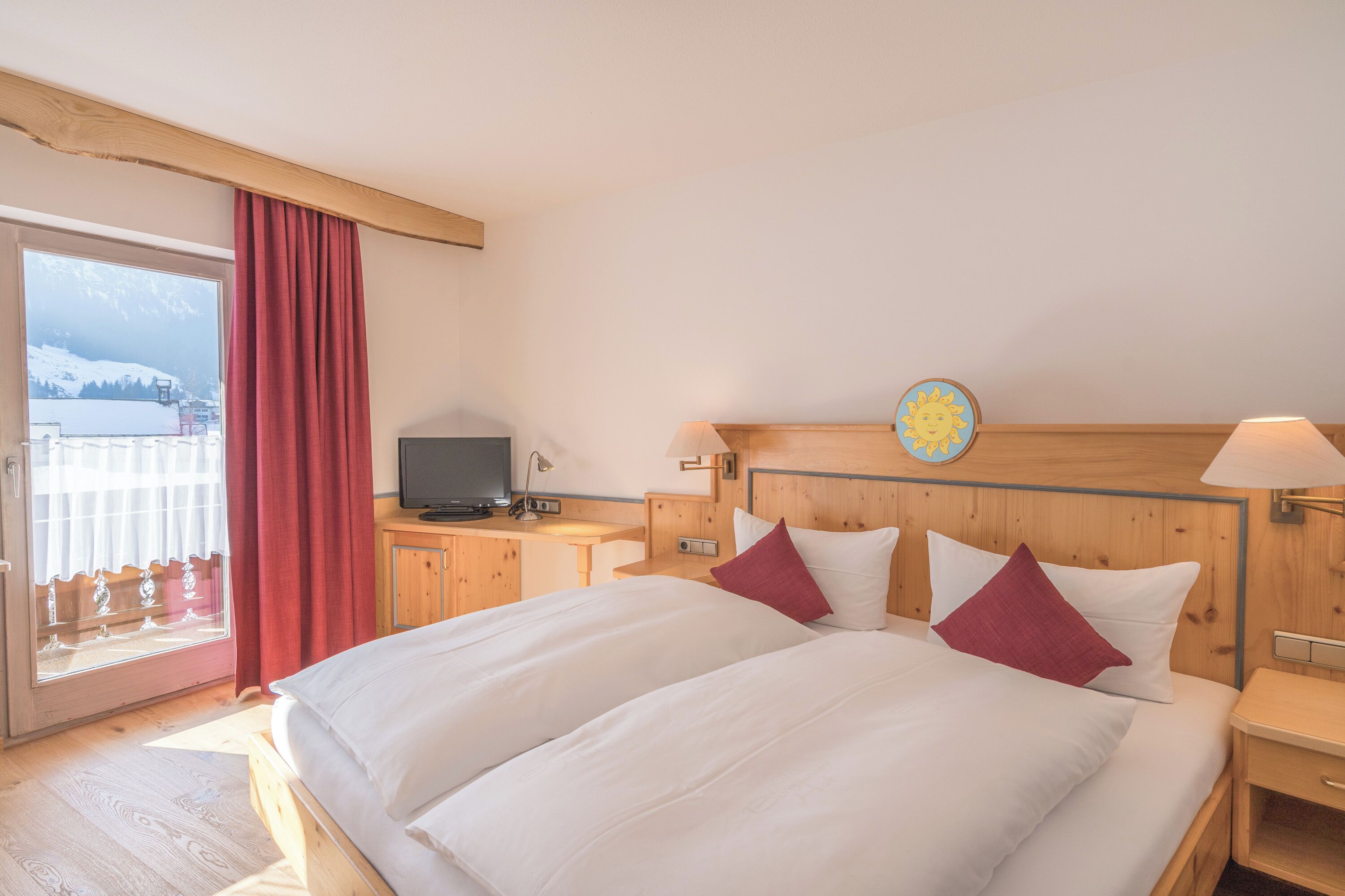 Habitación de hotel con una cama grande, cabecera de madera, un televisor sobre una mesa de madera y vistas a las montañas a través de una ventana.