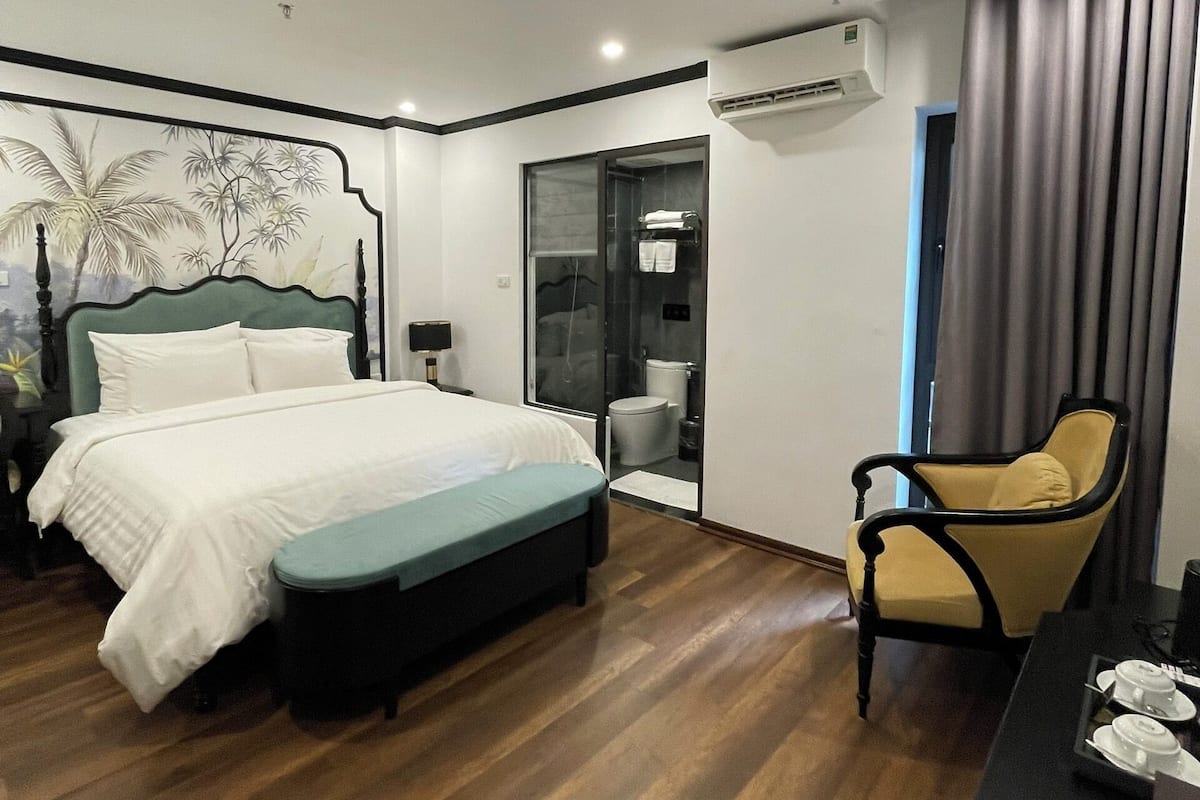 A modern hotel room with a large bed, a small seating area, a desk with a chair, and a bathroom visible through a glass door.