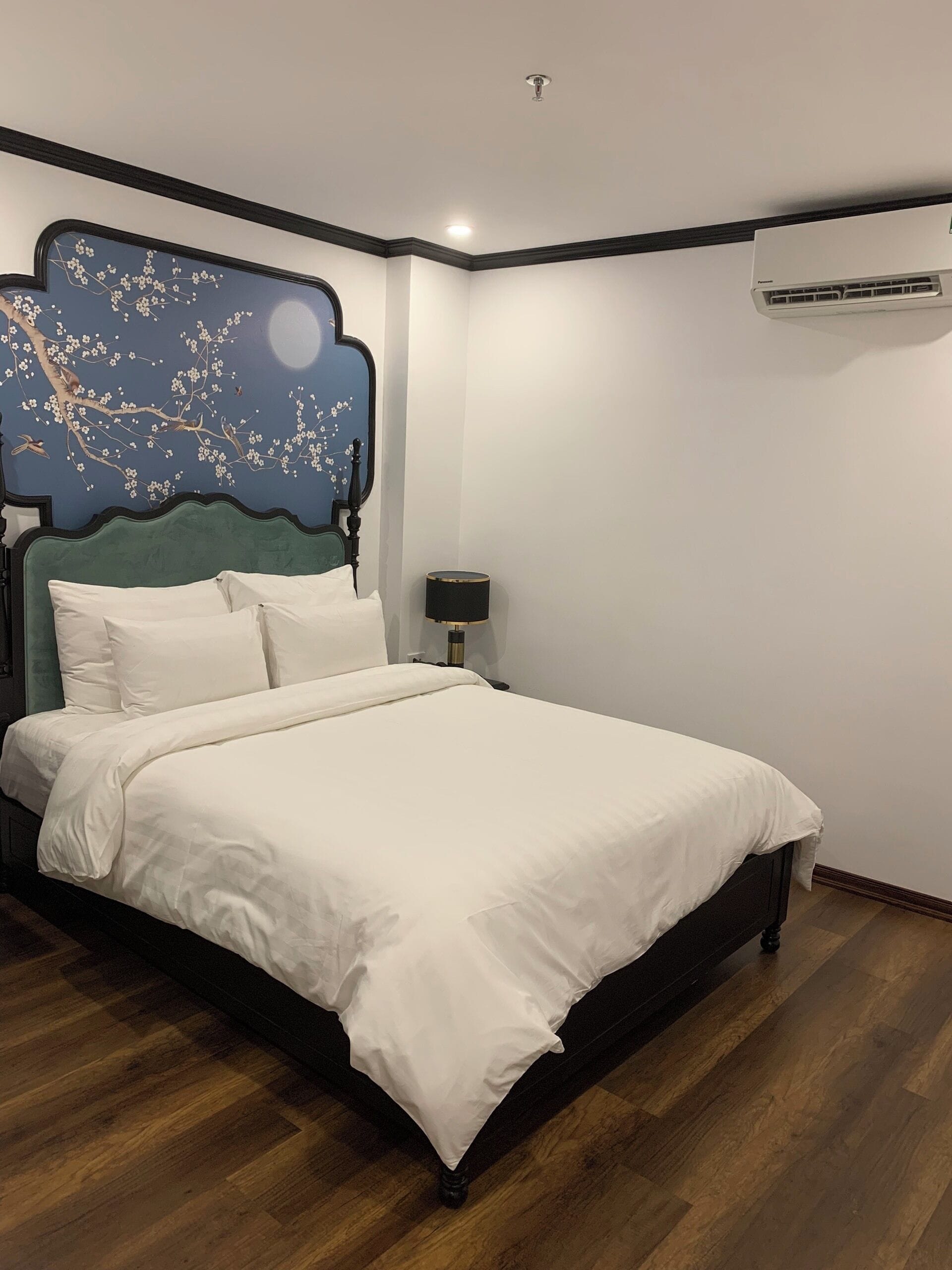 A neatly made bed with white linens in a room with a dark wooden floor, a wall-mounted air conditioner, and a decorative wall piece.