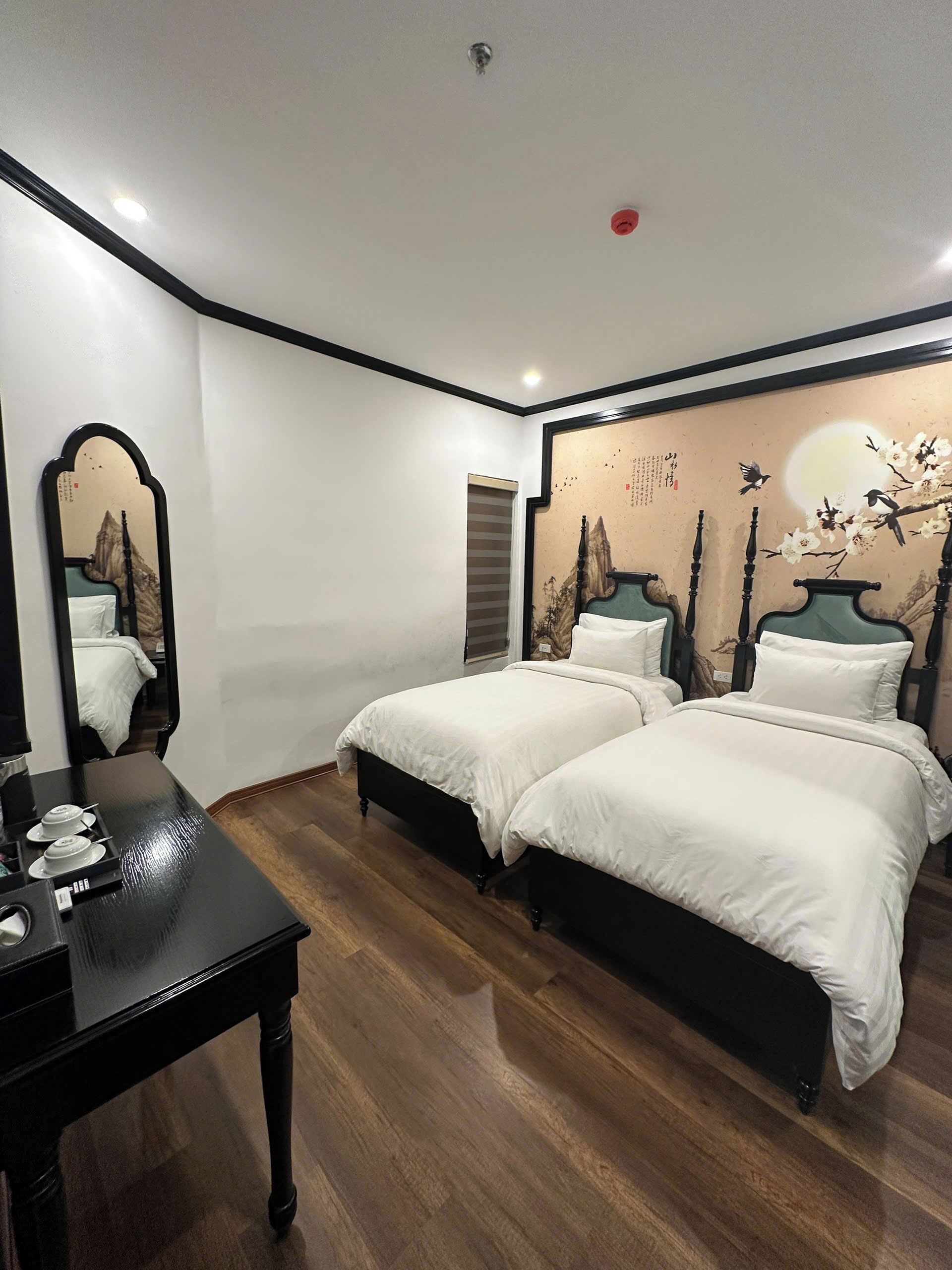 A bedroom with two beds, a desk, a mirror, and a wall with a painting.