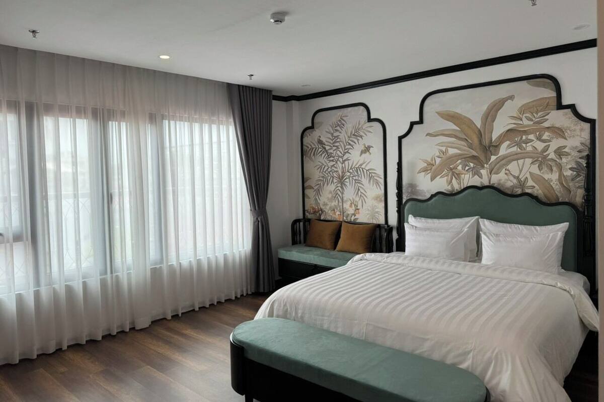 A hotel room with a large bed, a bench, a window with sheer curtains, and a wall with a floral mural.