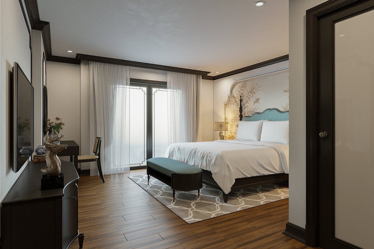 A modern hotel room with a wooden floor, a bed with white bedding, a desk with a vase, and a large window with sheer curtains.
