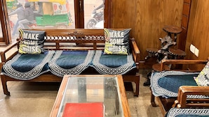 A room with a wooden sofa set, a glass-top table with a red book, and a view of the street through a large window. - Hotel Sunshine (Gurugram)