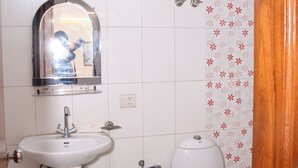 A small bathroom with a pedestal sink, a toilet, a blue bucket, and a water heater mounted on the wall. - Hotel Sunshine (Gurugram)