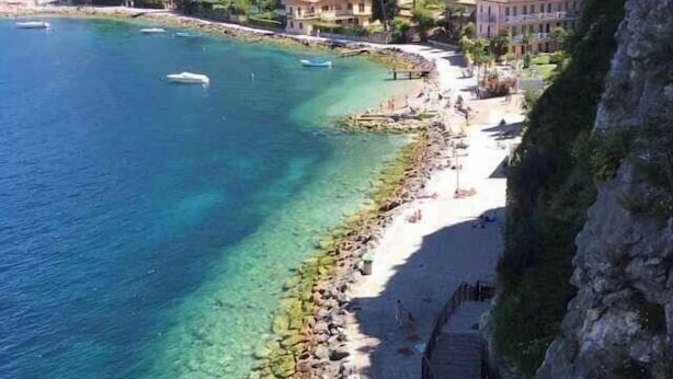 A coastal area with clear blue waters, a sandy beach, and a rocky shoreline. There are buildings along the coast, and a wooden structure is visible on the beach. - Villa Teresa-Lake View (Torri del Benaco)