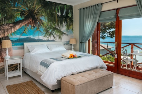 A bedroom with a bed, a nightstand, a lamp, a bench, and a view of the sea through a balcony.