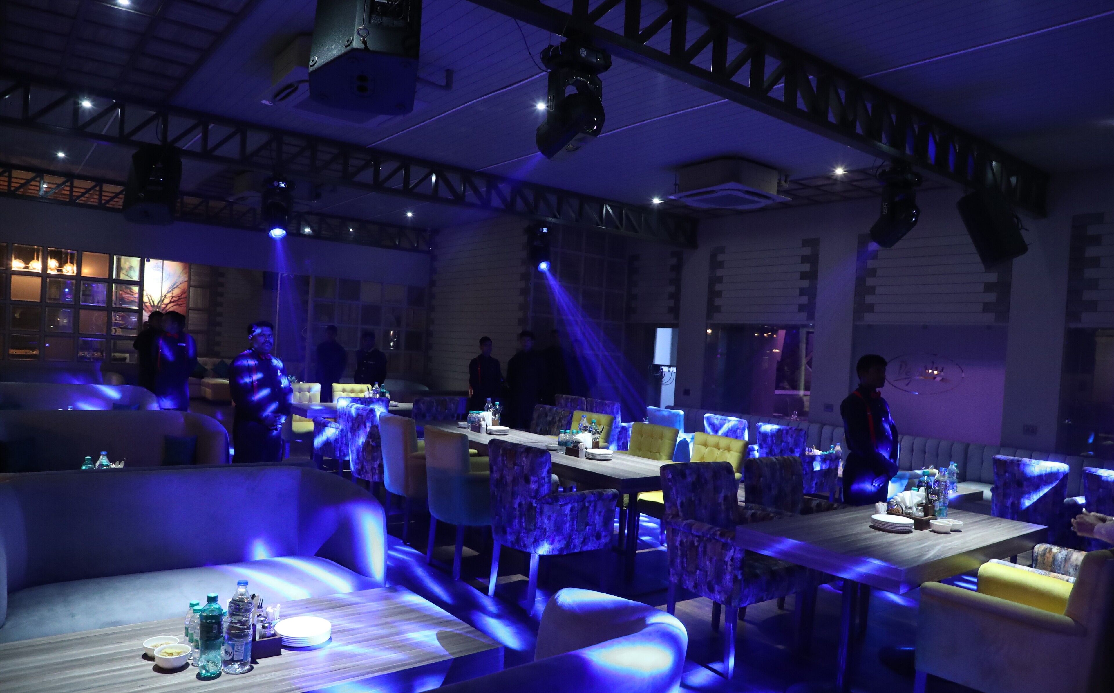 A restaurant with tables and chairs, some with blue lighting. There are people in the background.