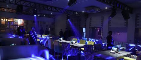 A restaurant with tables and chairs, some with blue lighting. There are people in the background.