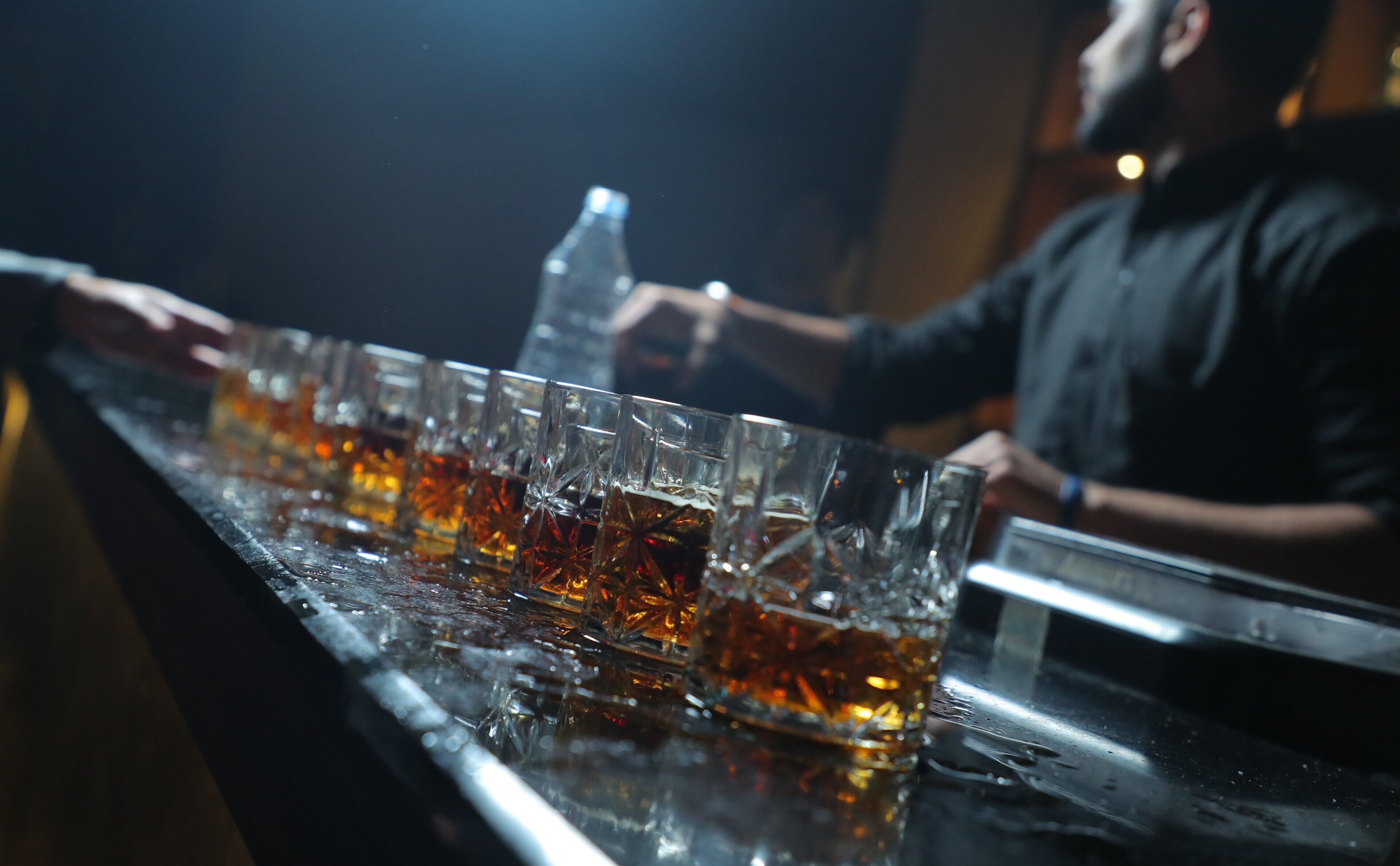 Una barra con varios vasos de líquido ámbar, probablemente un licor, y un bartender preparando bebidas.